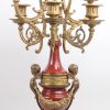 Een driedelig klokstel van verguld brons en roodgeglazuurd aardewerk, waarbij de pendule versierd met twee saters en de kandelaars met engelenkopjes.
