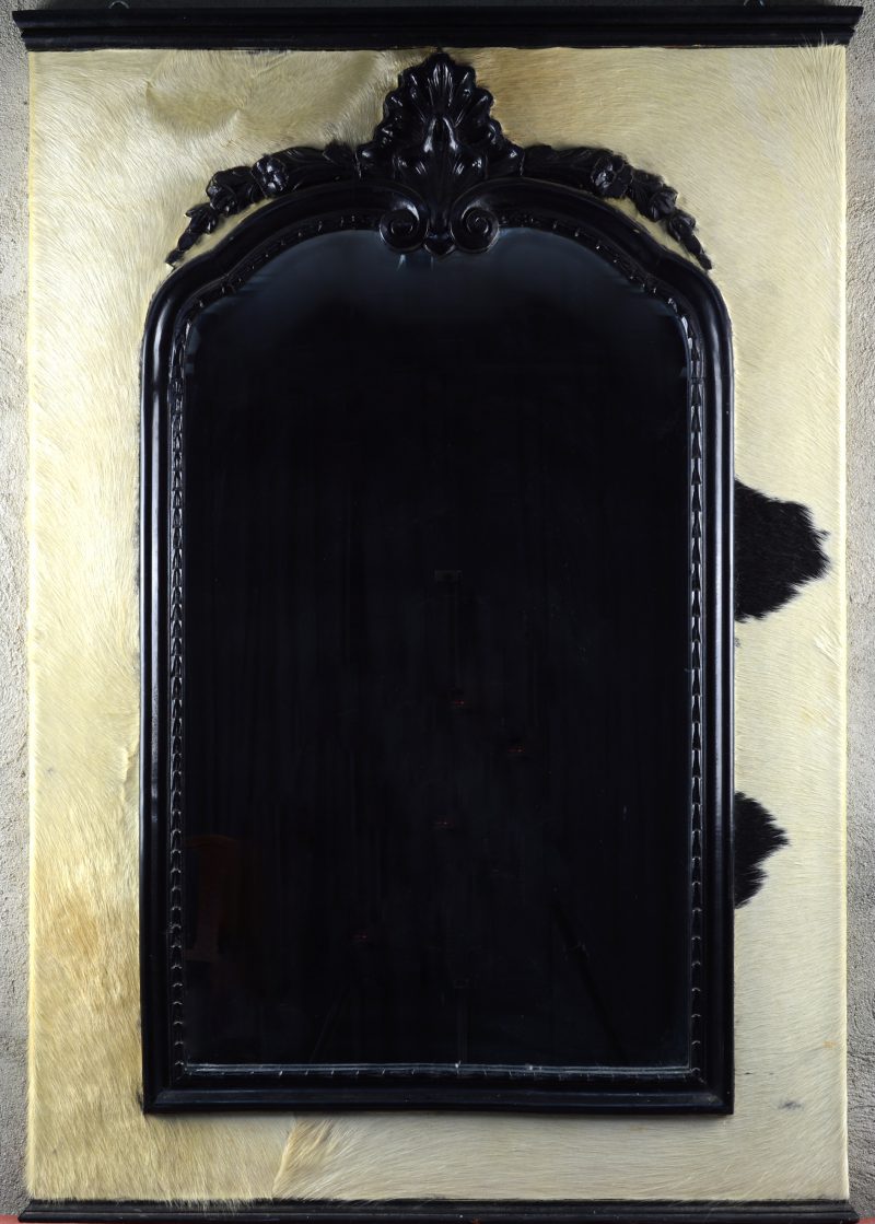 Een houten wandspiegel met zwart gepatineerd houten lijst en bekleed met koeienvacht.
