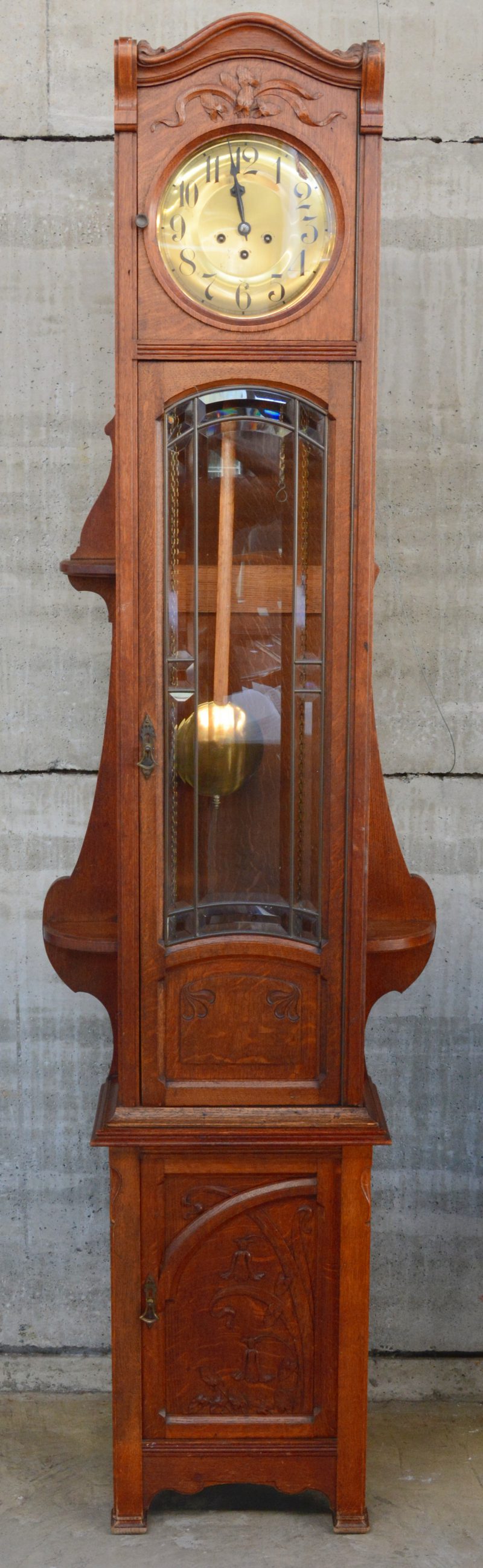 Een art nouveau staande klok met uitgestoken versieringen en met in facet geslepen glas in de deur.