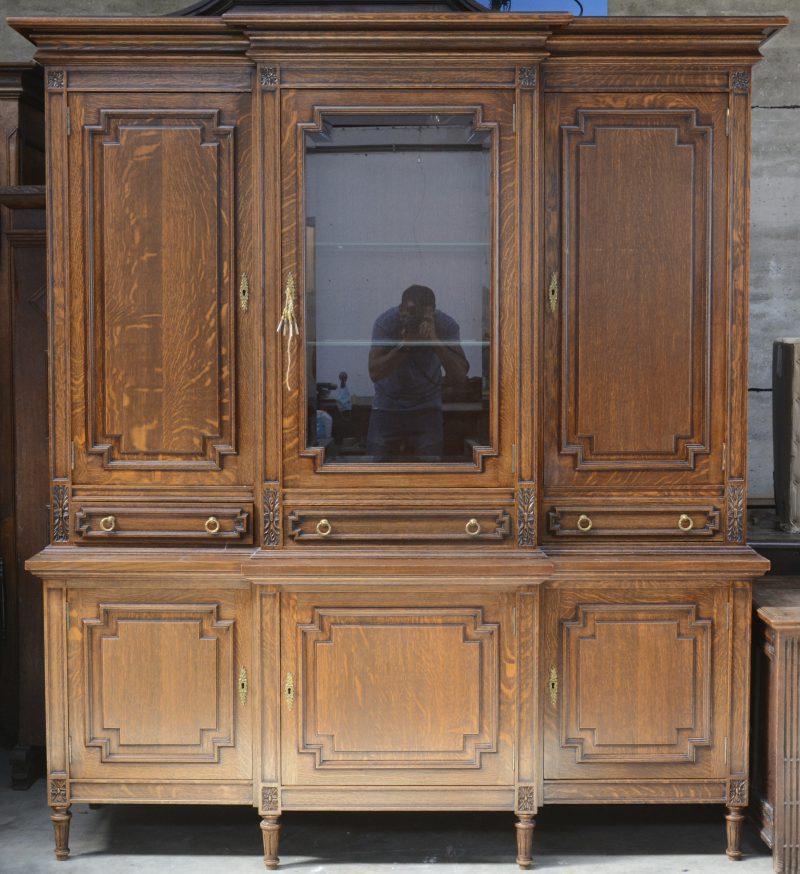 Een tweeledige eikenhouten breakfront buffet met onderaan drie paneeldeuren en bovenaan twee paneeldeuren aan weerszijden van een gevitreerde deur en drie laden. Lodewijk XVI-stijl. In 1992 op bestelling gemaakt voor 760.040 BEF (op factuur).