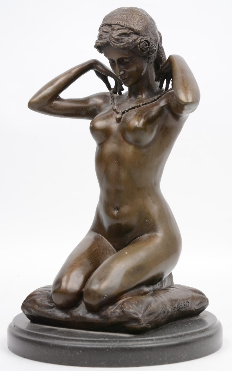 “Vrouwelijk naakt met hanger, knielend op een kussen”. Een bronzen beeld naar een werk van Paul Ponsard. Op zwart arduinen sokkel.
