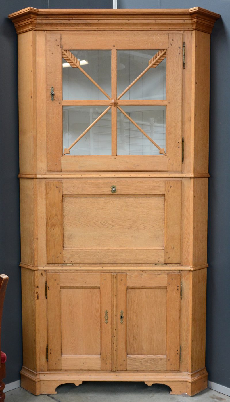 Een Gelderse hoekkast van blank eikenhout met twee paneeldeurtjes onderaan, een schijfblad en een gevitreerde deur bovenaan. Omstreeks 1820.
