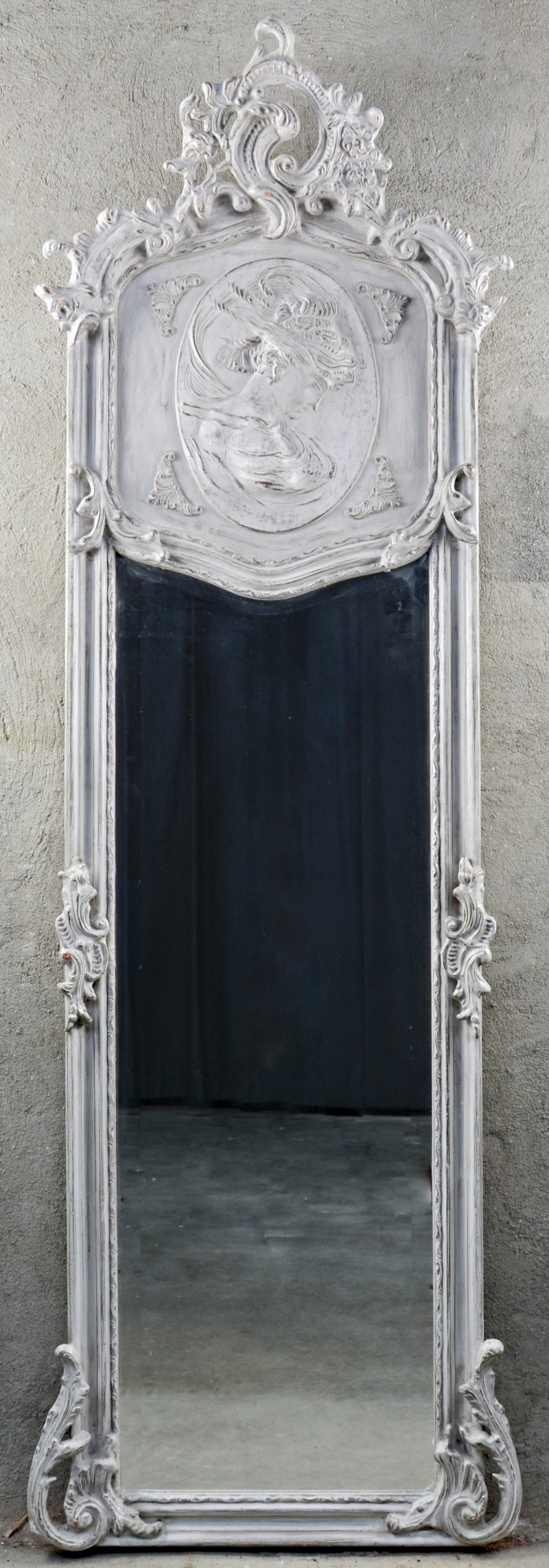 Een grijsgepatineerde houten spiegel in Lodewijk XV-stijl, versierd met een vrouwenprofiel in reliëf.