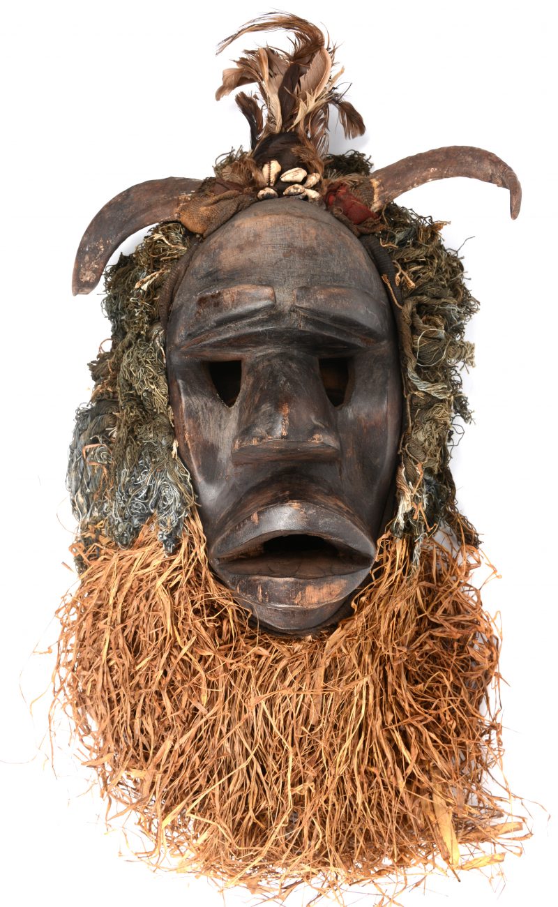 Een Afrikaans houten masker met wol, droog gras, veren en geitenhoorns.