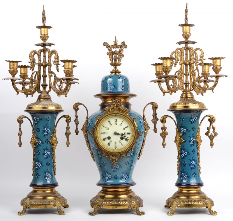 Een driedelig klokstel van koper en porselein met een blauw decor van korenbloemen. Begin XXste eeuw. Met sleutels en slinger.