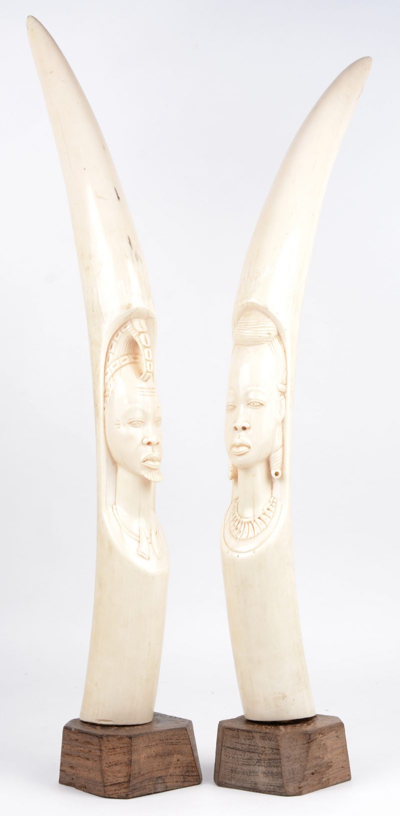 Een paar bewerkte Afrikaanse slagtanden met voorstelling van een mannen- en een vrouwenhoofd. Op houten sokkel. Pré 1947.