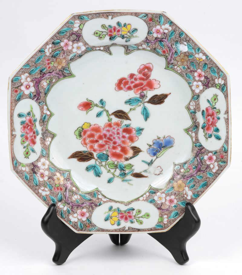 Een achthoekig bord van Chinees porselein met een famille rose decor van pioenen op het plat en met bloemen in en rond uitsparingen op de vleugel. Tijdperk Qinglong.