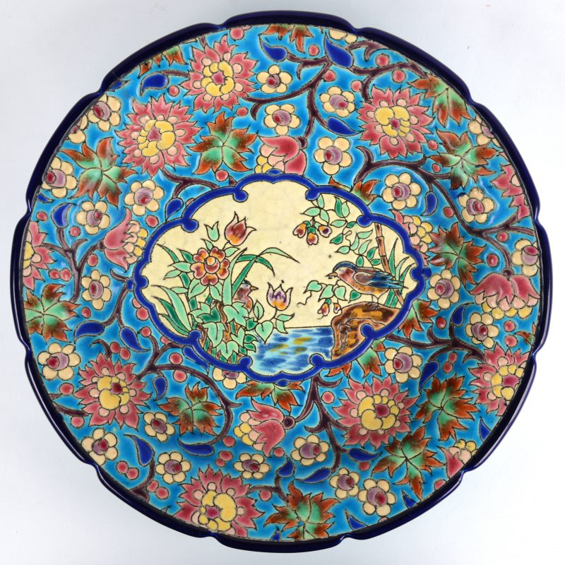 Een diepe ronde schaal van meerkkleurig aardewerk, versierd met een decor van vogels bij een vijver in een cartouche in het plat, omgeven door diverse bloemen. Onderaan gemerkt. Haarscheurtje.
