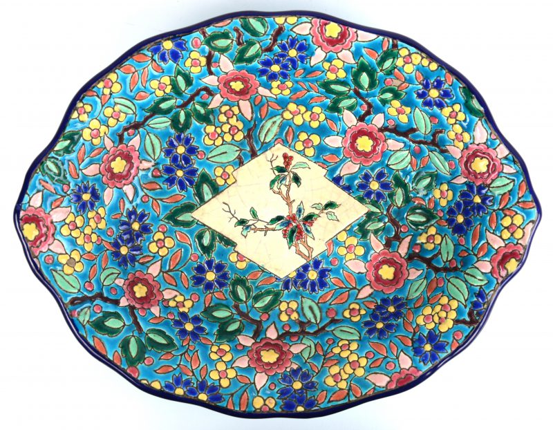 Een diepe ovale schaal van meerkkleurig aardewerk, versierd met een decor van een plant in een cartouche in het plat, omgeven door diverse bloemen. Onderaan gemerkt. Haarscheurtje.