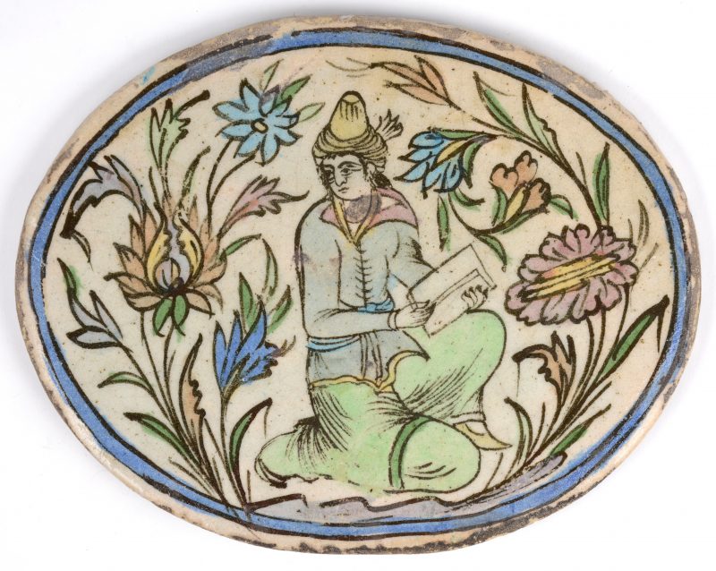 Een ovale plaquette van Perzisch aardewerk met een meerkleurige afbeelding vn een schrijver.