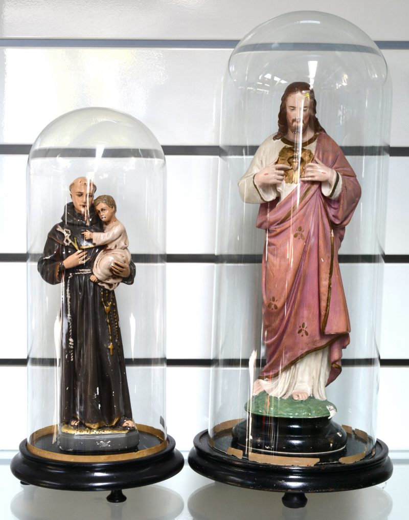 “Sint Antonius” & “Christus Heilig Hart”. Twee gepolychromeerde plaasteren Heiligenbeelden onder stolp. (De tweede met schilfer aan de voet).
