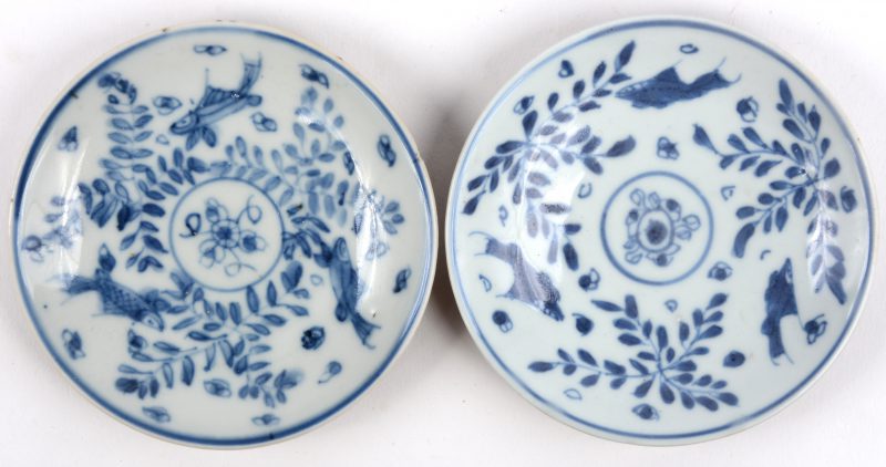 Een paar schoteltjes van Chinees porselein met een blauw en wit decor van vissen en met capucineporselein op de onderkant. Eén met zeer kleine randschilfer.