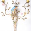 Een paar kandelaars van meerkleurig porselein en messing, opgebouwd uit een sierstuk, getooid met een papegaai en met twee lichtpunten in de vorm van plantenranken met porseleinen bloemen. Apocrief gemerkt achteraan.