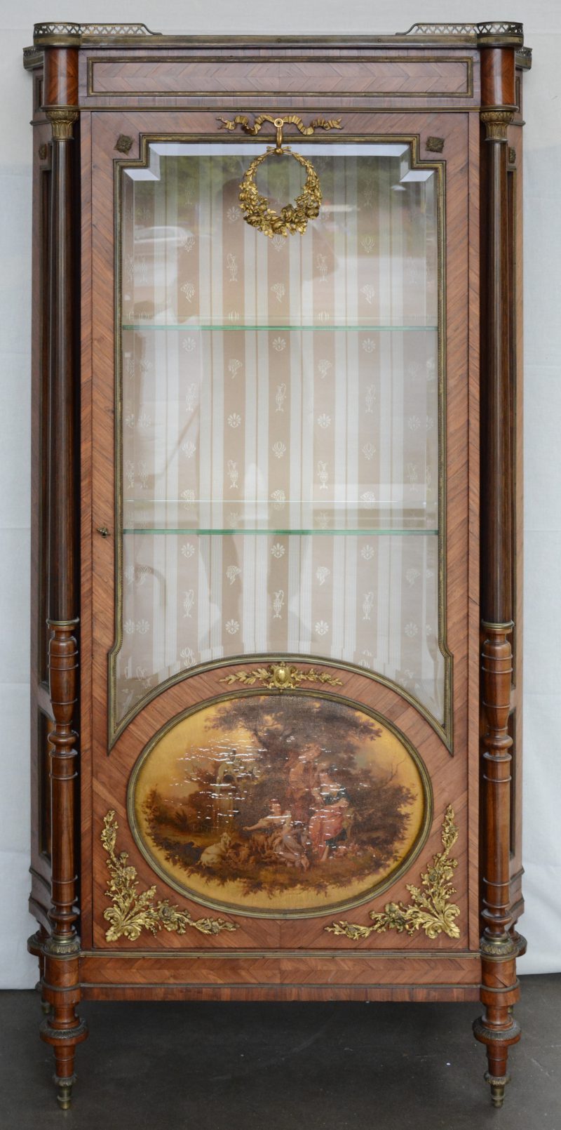 Een gefineerde ééndeursvitrine in Lodewijk XVI-stijl met een ovale schildering in de deur en het geheel versierd met verguld beslag. Slijtage aan de schildering.