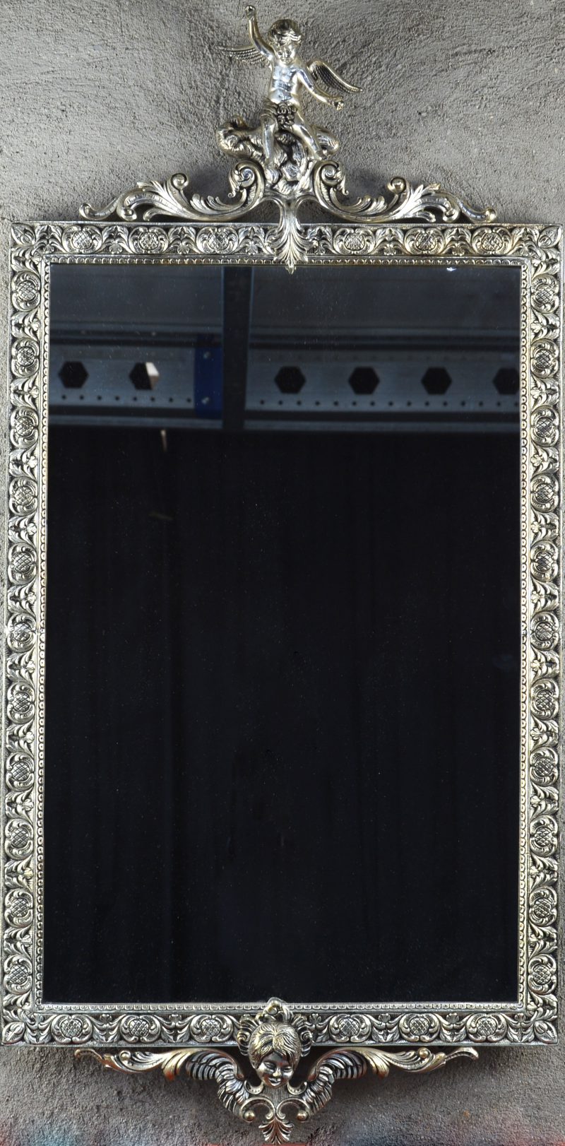 Een spiegel in verzilverd metalen lijst, getooid met een engeltje bovenaan en met engelenkopje onderaan.