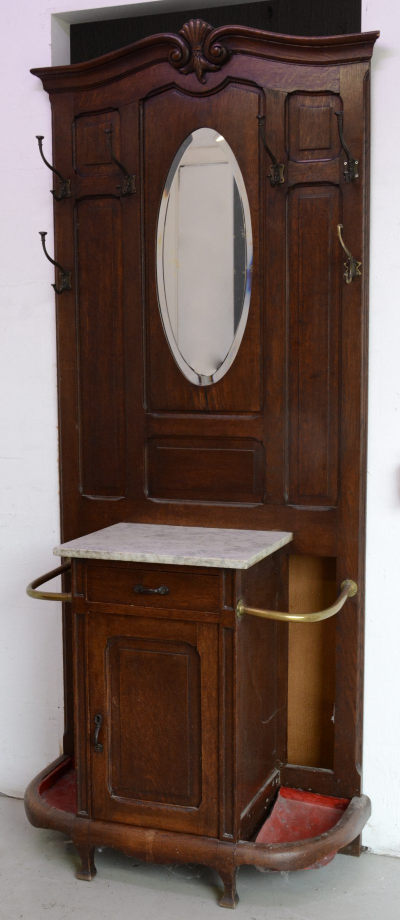 Een grote wandkapstok met spiegeltje en aan weerskanten een paraplustaander met metalen bodem. Centraal een deurtje en een lade.