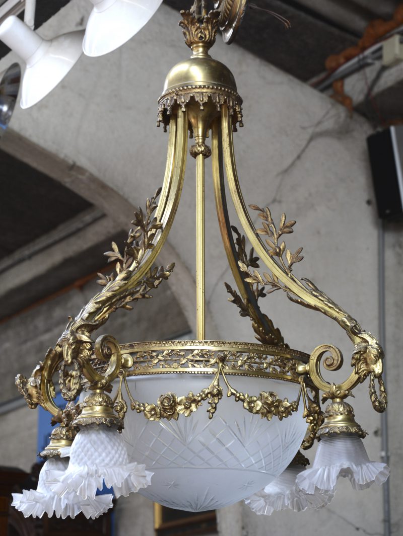 Een verguld bronzen luchter met vier lichtarmen, versierd met ramskoppen en met gesatineerde glazen kapjes en geslepen centrale coupe.