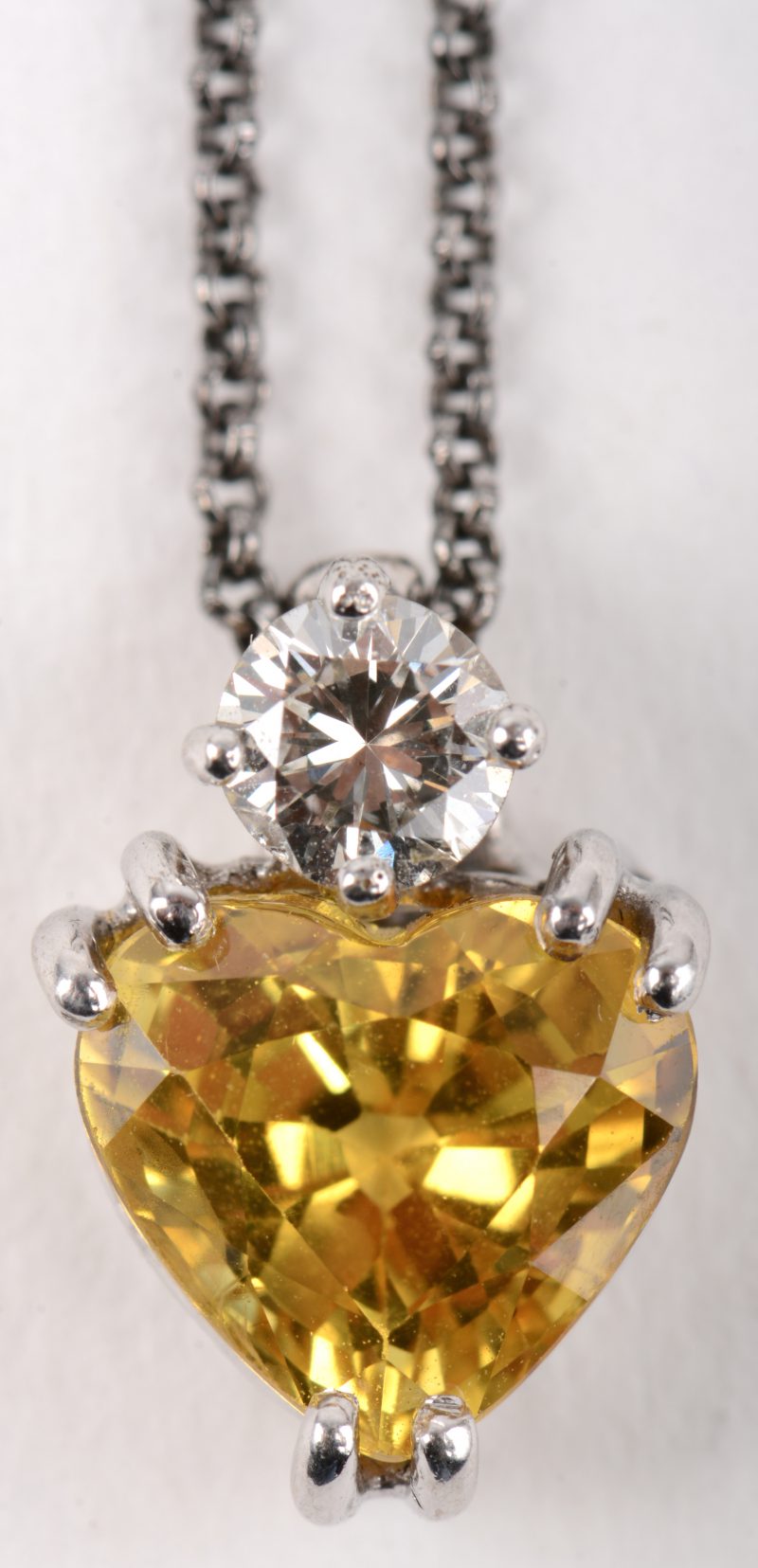Een 18 K grijskleurige gouden ketting met hartvormige hanger van 18 K witgoud bezet met een briljant van +- 0,20 ct. en een gele saffier van +- 2,15 ct.