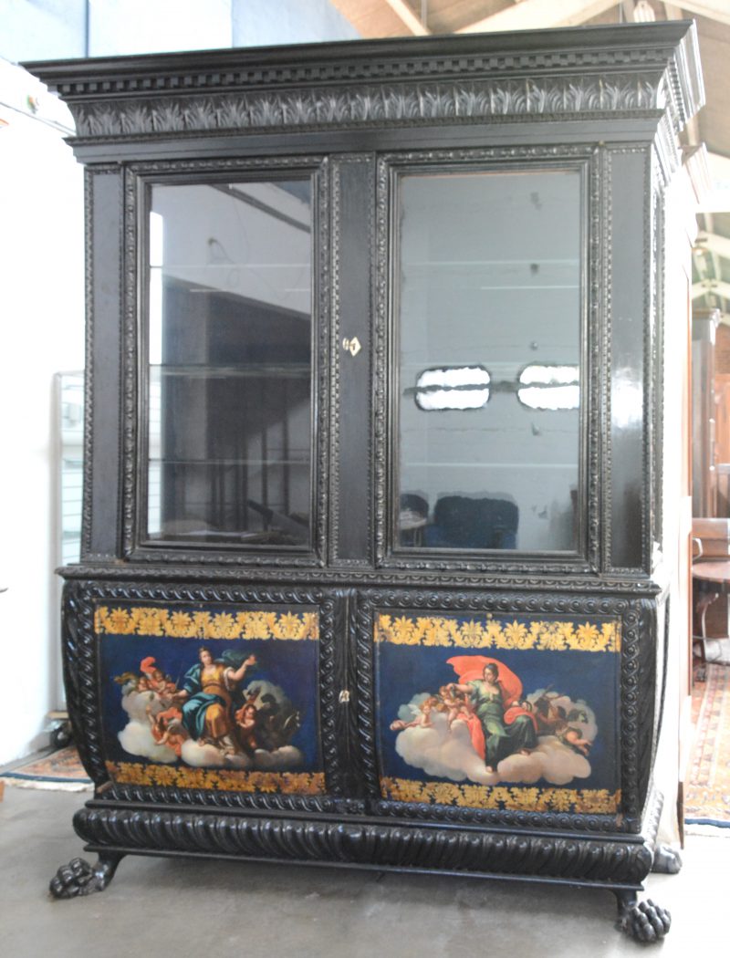 Een vitrinekast van gebeeldhouwd en zwartgepatineerd hout  op eeuwenpoten. Onderaan twee gebombeerde paneeldeuren, versierd met handgeschilderde allegorische decors, omzoomd met acanthuisbladeren. Tijdperk Napoleon III.