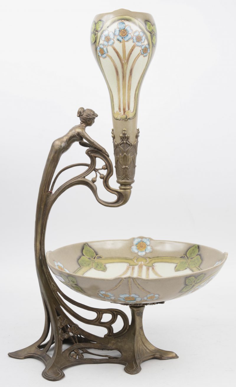Een sierstuk in art-nouveaustijl met een aardewerken schotel onder een vaas, versierd met een meerkleurig bloemendecor in bronzen staander, versierd met een vrouwenfiguur.