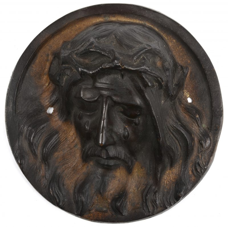 Een ronde bronzen plaquette met voorstelling van het hoofd van Christus met de doornkroon. Achteraan gesigneerd ‘S. Norca’.