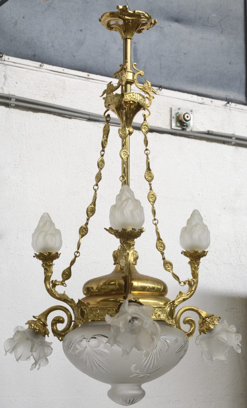 Een verguld bronzen luchter met drie lichtarmen, eindigend in bloemvormige kapjes, drie opstaande lichtpunten met vlamvormige kapjes en met een centrale coupes.