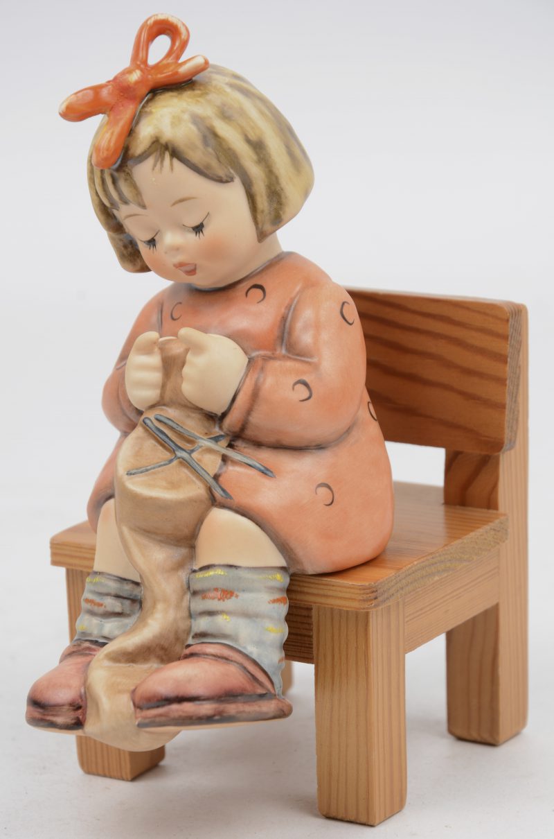 “To keep you warm”. Een beeldje van meerkleurig biscuit uit de reeks van M.I. Hummel. Onderaan gemerkt. Op houten stoeltje.