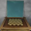 Een gefineerde notenhouten speeltafel met uitneembaar speelvlak met aan de ene kant fineer, aan de andere groen vilt en eronder een schaakbord. Onder het kantelblad een opbergruimte.