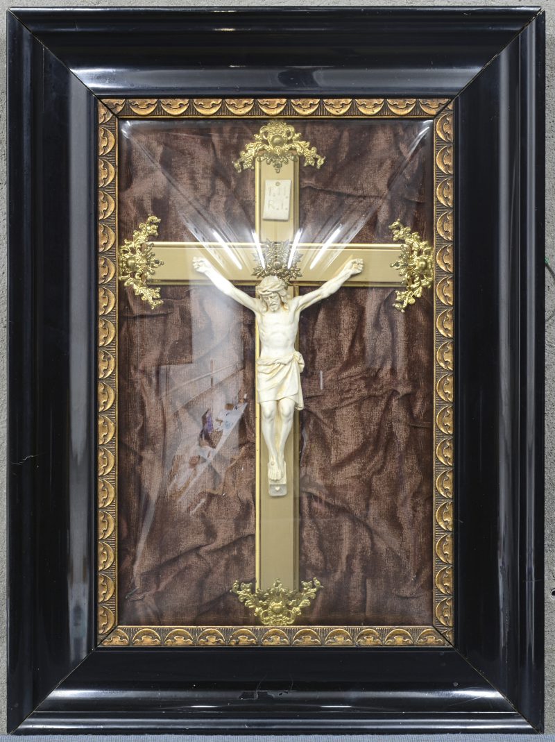 Een messingen kruis met gipsen(?) Christus. Achter bol glas en in zwartgelakt houten kader.
