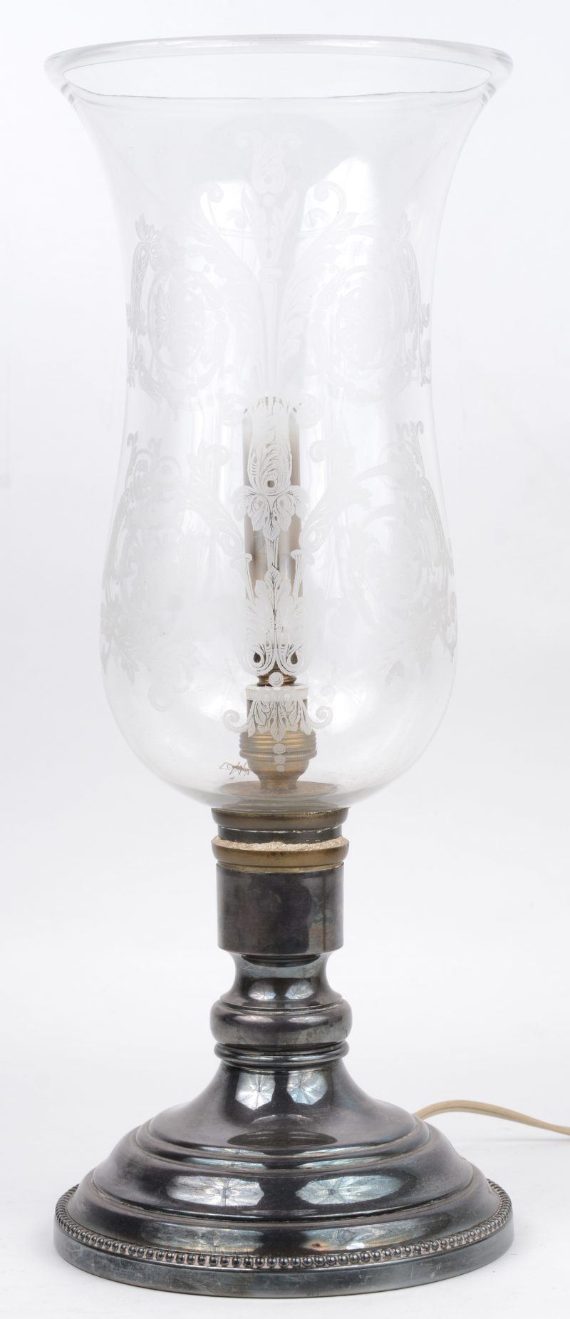 Een windlamp op verzilverde voet met gegraveerde glazen kap. Op elektriciteit.