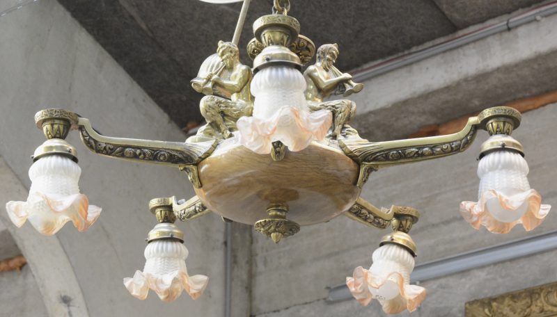 Een luchter van verguld brons en hout met vijf lichtpunten, versierd met fluitspelende saters en eindigend in bloemvormige glazen kapjes.