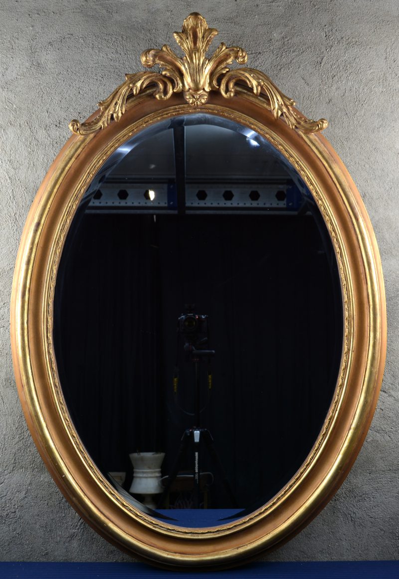 Een ovale spiegel van goudgepatineerd hout met een acanthusblad in de kuif.