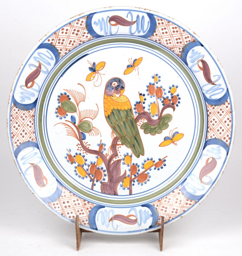 Een aardewerken sierschotel met een meerkleurig decor van een papegaai en vlinders bij een bloeiende tak. Achteraan gemerkt ‘B.D.’. Omstreeks 1800.