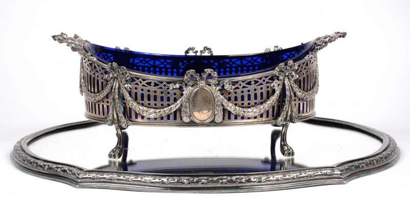Een zilveren milieu-de-table, versierd met strikken en guirlandes, gemerkt met Engelse keuren. Met blauw glazen recipiënt. Op verzilverd metalen spiegel.