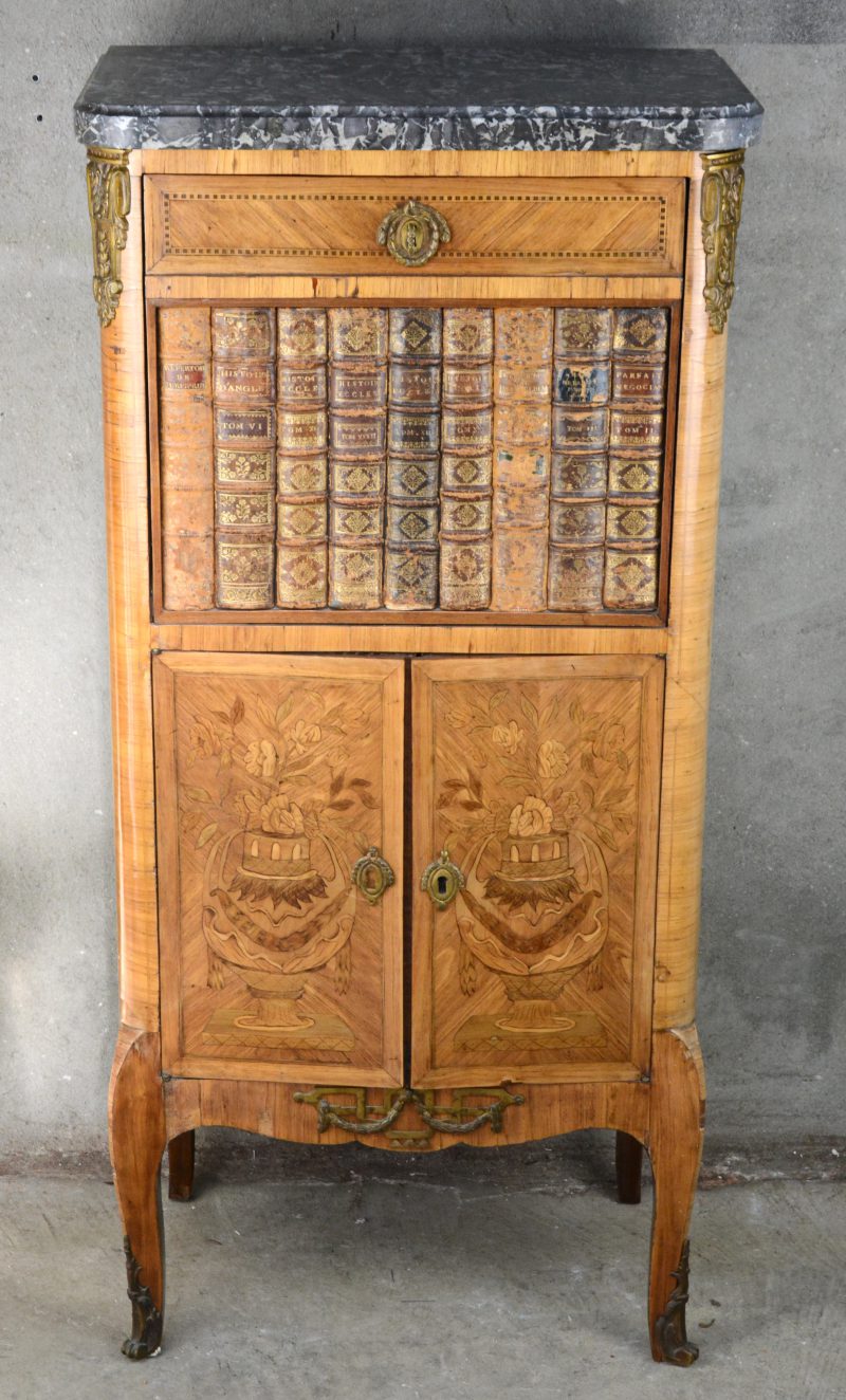 Een klein secretaire van gefineerd hout in Régencestijl versierd met siervazen van marqueterie met een klapblad met boekenruggen in trompe l’oeil.