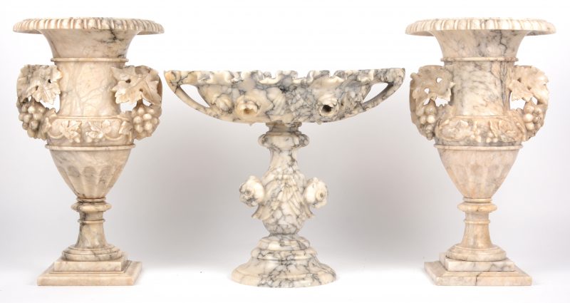 Een driedelig kaststel van gesculpteerd albast, bestaande uit twee siervazen en een coupe op voet. Versieringen van druivenraken en bloemen.