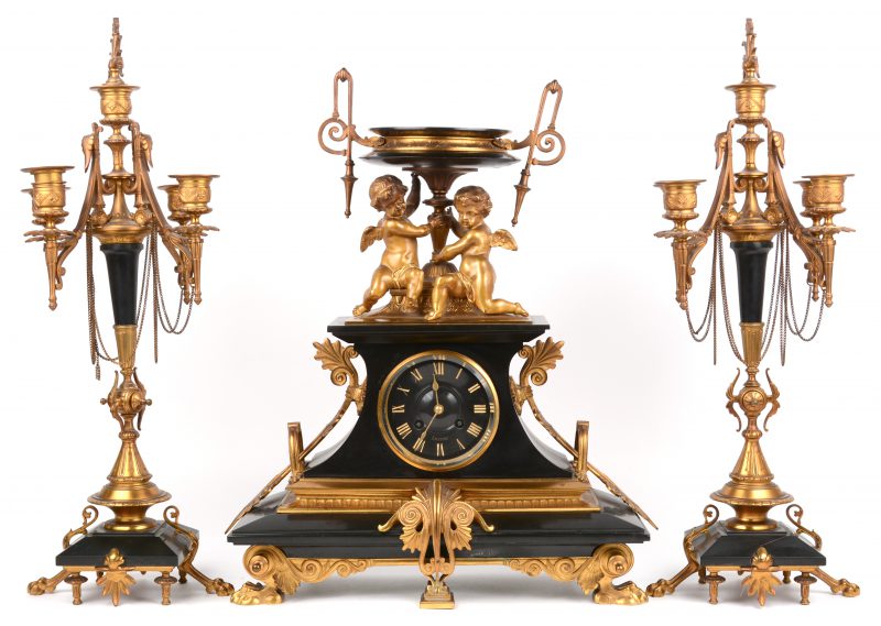 Een driedelig schouwgarnituur van verguld brons en arduin, bestaande uit een pendule, versierd met engeltjes en twee kandelaars met vijf lichtpunten. Eind XIXe eeuw. Kleine beschadiging bovenaan.