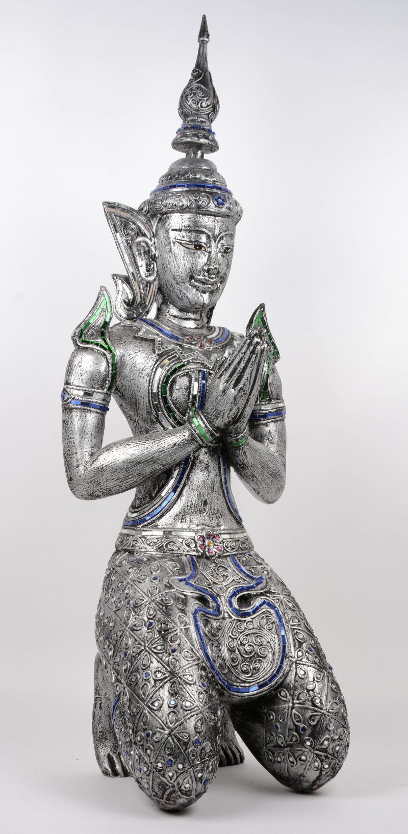 Een knielende Thaise Boeddha van zilvergepatineerd hout, versierd met glazen plaatjes in groen, rood en blauw.