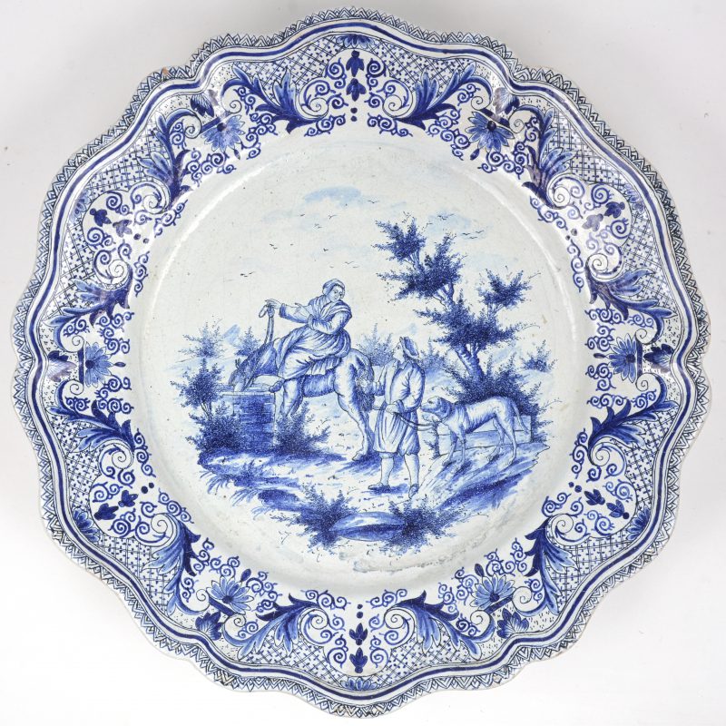 Een grote gelobde schotel van aardewerk met blauw op wit decor van personages in een landschap. Gemerkt op de verso.