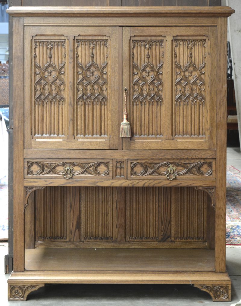 Een eikenhouten dressoir/ televisiemeubel in neogotische stijl met twee deels naar binnen openende deurtjes versierd met briefpaneel. Binnenin een uitschuifbaar blad.