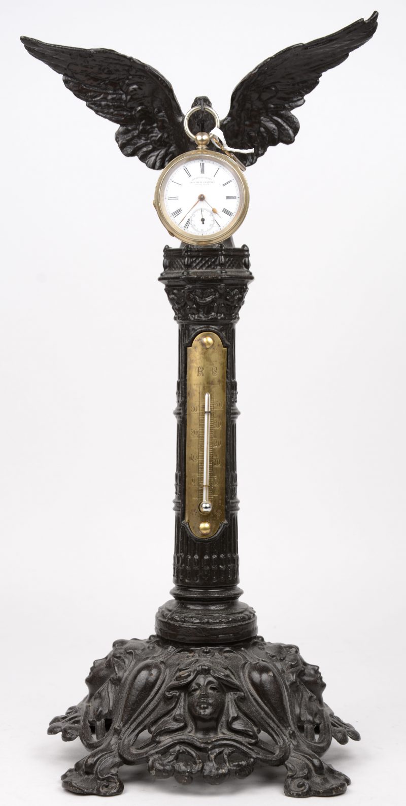 Een gepatineerd zamakken horlogehouder in de vorm van een zuiltje, versierd met vrouwenhoofden in de basis, met thermometer op de kolom en bovenaan getooid met een adelaar. We voegen er een Zwitsers zakhorloge in verzilverd metalen kast aan toe. Begin XXe eeuw.