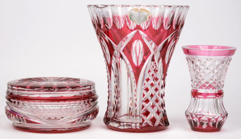Een lot geslepen en rood gedubbeld kristal, bestaande uit een bonbonière en een grote en een kleine vaas. Allen gemerkt onderaan. De grootste vaas met beschadiging aan de rand.