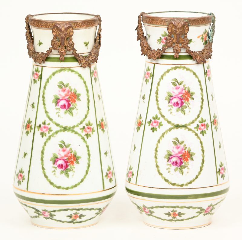 Twee opconische porseleinen siervaasjes, versierd met meerkleurig bloemendecor en met koperen monturen met guirlandes en ramskoppen. Gemerkt onderaan “Versailles” onder een groene adelaar.