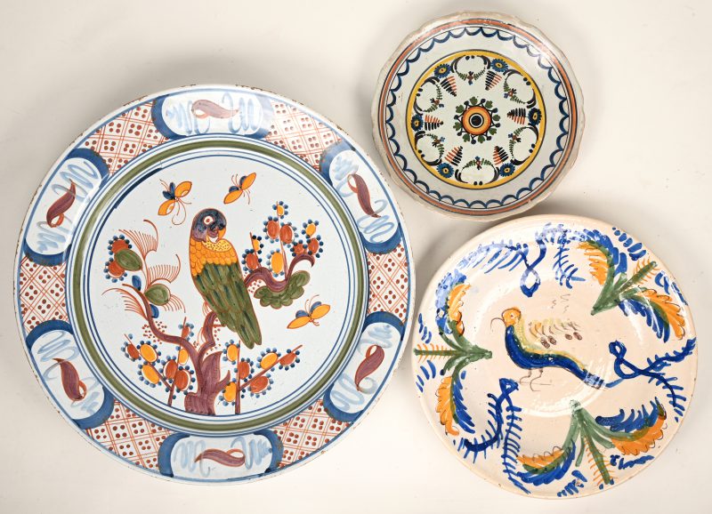 Een aardewerken sierschotel met een meerkleurig decor van een papegaai en vlinders bij een bloeiende tak. Achteraan gemerkt ‘B.D.’. Omstreeks 1800. En twee andere.