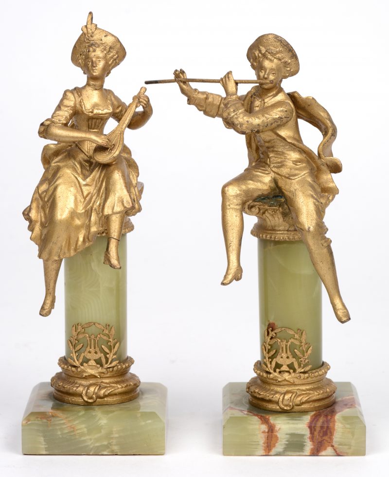 Twee kleine vergulde metalen beeldjes van een jong musicerend koppel gezeten op onyxen zuiltjes met vergulde ionische kapitelen. XIXde eeuw.
