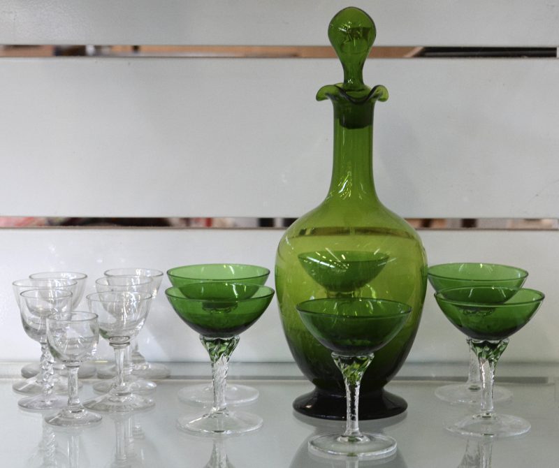 Een karafje van groen glas met zes bijpassende glaasjes. We voegen er zeven kleurloze borrelglaasjes, waaronder enkele van Elixir d’Anvers aan toe.