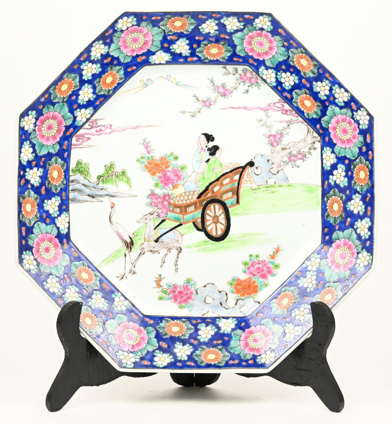Een achthoekige schotel van meerkleurig chinees porselein met bloemen, een reiger en twee vrouwen in een riksja voortgetrokken door een ree in het decor.