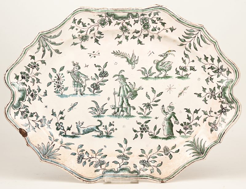 Langwerpige schotel van aardewerk met een groen en sepia decor van personages, dieren en bloemen en met gechantourneerde rand. XVIIIe eeuw. Onderaan gemerkt. Een randschilfer.