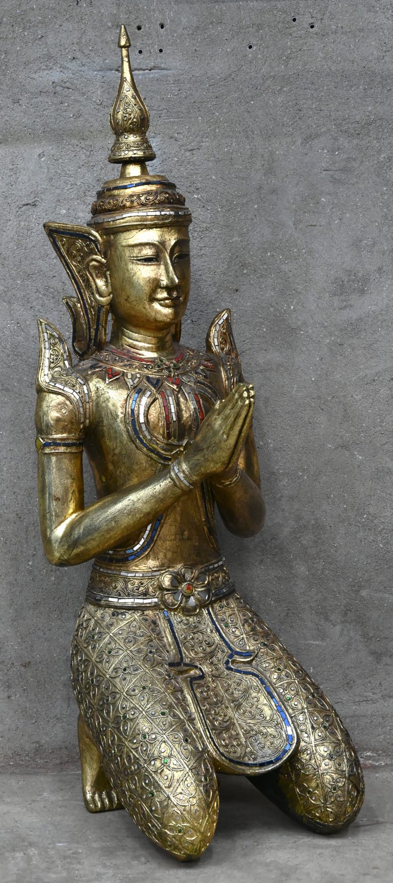 Een knielende Thaise Boeddha van verguld hout, versierd met gekleurd glazen elementen.