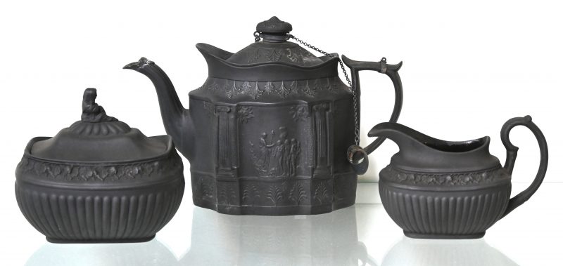 Een theeset van basaltware bestaande uit een theepot met classicistisch decor, suikerpot en melkkannetje met druivenranken en een zittende figuur boven aan het deksel.